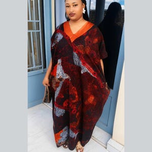 May include: A floor-length kaftan dress featuring a V-neckline with a solid orange trim. The fabric displays a dark, abstract pattern in shades of deep red, black, and grey, with distinct circular motifs and textured lines across the loose-fitting garment.