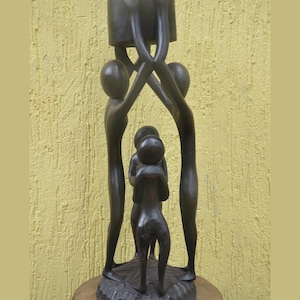 May include: A dark brown wooden sculpture featuring four stylized figures standing on a circular base. Two taller figures stand on the outer edges, their arms raised to support a cylindrical platform above, while two smaller figures stand in the center.