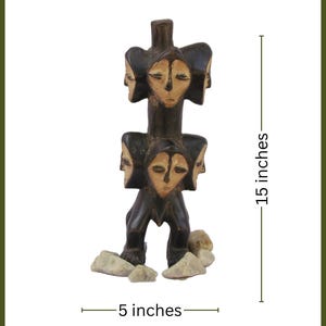 May include: A dark brown wooden sculpture with multiple carved faces. The sculpture is 38 cm tall and 13 cm wide, with a weight of 4.1 kg. The base includes several small, light-coloured stones. The text "KIVU HOME" is in the upper right corner.