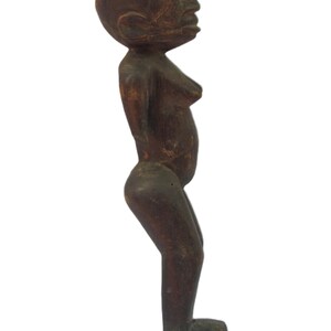 以下が含まれることがあります： 立っている人物の木彫りの置物。濃い茶色で木目がはっきりと見え、丸い頭、くっきりとした顔立ち、わずかに突き出た腹部が特徴です。人物の腕は体の両側にあります。