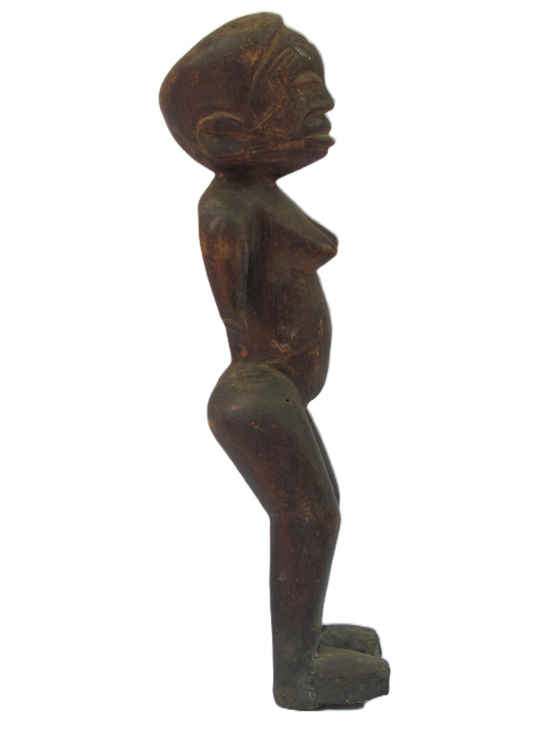 以下が含まれることがあります： 立っている人物の木彫りの置物。濃い茶色で木目がはっきりと見え、丸い頭、くっきりとした顔立ち、わずかに突き出た腹部が特徴です。人物の腕は体の両側にあります。