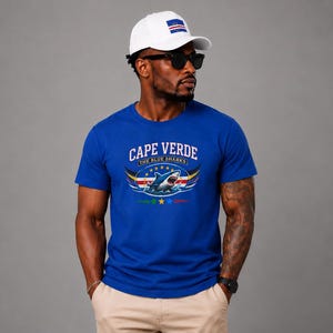 May include: A royal blue short-sleeved t-shirt featuring a graphic of a shark with the text CAPE VERDE THE BLUE SHARKS and a row of five stars. The shirt is paired with a white baseball cap displaying the Cape Verde flag on the front panel.