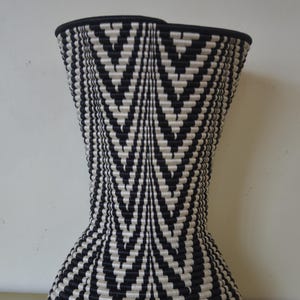 May include: A decorative woven stool featuring a black and white chevron pattern. The stool has a flared base, a narrow waist, and a wider top. The top edge is finished with a black border. The design is symmetrical and geometric.