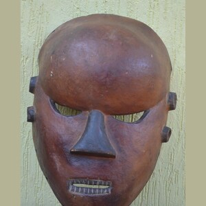 May include: A brown wooden mask with a smooth, rounded forehead, a triangular nose, and horizontal eye openings. The mask features a rectangular mouth with visible carved teeth and small circular protrusions on both sides of the face.