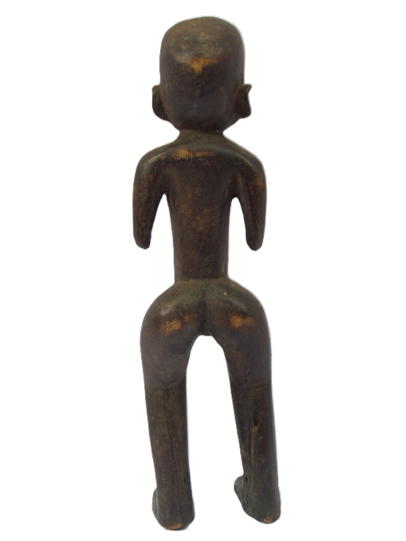 以下が含まれることがあります： 暗い茶色の木製の置物で、様式化された人間の形を描いています。 このフィギュアは丸い頭、小さな耳、腕を広げています。 脚は分離しており、足が底にあります。 彫刻は風化した外観をしています。
