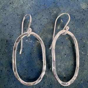 May include: A pair of silver oval earrings with a hammered texture. The earrings have a simple hook design and are displayed against a blue background. The earrings are approximately 1.5 inches in length.