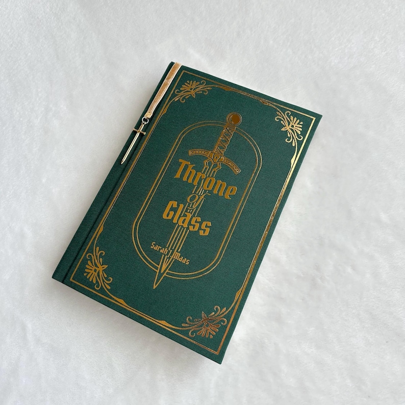 K&ouml;nnte beinhalten: Ein gr&uuml;nes Hardcover-Buch mit goldenen Lettern und einem goldenen Schwertdesign. Der Titel des Buches lautet "Throne of Glass" von Sarah J. Maas.