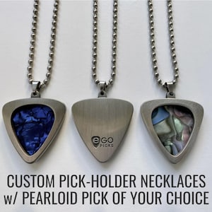 Puede incluir: Tres collares con soporte para púas de guitarra con cadenas de bolas. Los colgantes son plateados, triangulares y presentan una púa azul, iridiscente y un dorso liso. El texto en la parte inferior dice "CUSTOM PICK-HOLDER NECKLACES w/ PEARLOID PICK OF YOUR CHOICE."