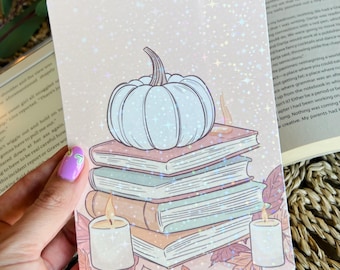 May include: A rectangular card with a light pink background featuring a stack of books, a white pumpkin, candles, and autumn leaves. The card has a glittery effect. The text on the card is partially visible.