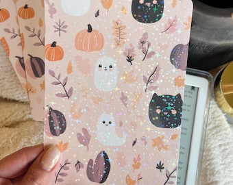May include: Kindle covers with a seasonal autumn design. The covers display pumpkins, ghosts, cats, and leaves in orange, purple, and white on a pink backdrop. The covers have a glittery, holographic finish.