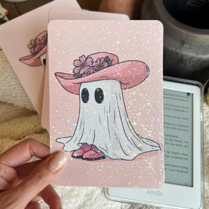 May include: A pink, glittery art print featuring a cartoon ghost wearing a pink hat adorned with flowers and pink shoes. The ghost has large, oval eyes. The background is a light pink with glitter accents. A hand holds the print.