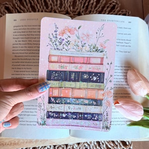 Könnte beinhalten: Eine dekorative Karte mit einem Stapel Bücher mit floralen Akzenten. Die Bücher haben verschiedene Farben und florale Designs. Die Karte liegt auf einem offenen Buch mit Text und einem gewebten Hintergrund.