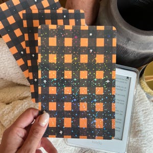 Könnte beinhalten: Mehrere rechteckige Karten mit einem schwarz-orangen Karomuster und holografischem Glitzer. Die Karten werden in der Nähe eines Kindle-eReaders gehalten. Die Karten haben ein Halloween-Thema.