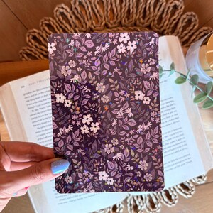 Könnte beinhalten: Eine rechteckige Karte mit einem floralen Muster in Lila-, Weiß- und Silbertönen. Das Design zeigt kleine Blumen und Blätter. Die Karte wird in einer Hand gehalten, mit einem Buch und dekorativen Elementen im Hintergrund.