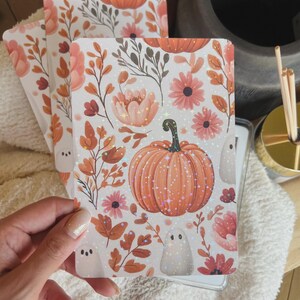 May include: A set of three rectangular cards with a fall-themed design. The cards feature a large orange pumpkin, pink flowers, and small white ghosts on a white background. The design includes orange and brown leaves and stems. The cards are held in a person's hand.