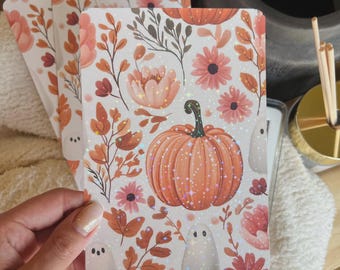 May include: Three rectangular cards with a seasonal autumn design. The cards feature an orange pumpkin, pink flowers, and small white ghosts on a white background. The design includes orange and brown leaves and stems. The cards are held in a person's hand.