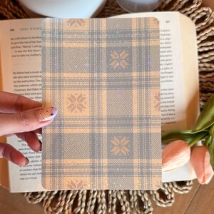 Peut inclure: Une carte rectangulaire à motif de carreaux gris et pêche avec des motifs de flocons de neige. La carte est tenue au-dessus d'un livre ouvert et d'un dessous de plat tressé, avec une bougie et des fleurs en arrière-plan.