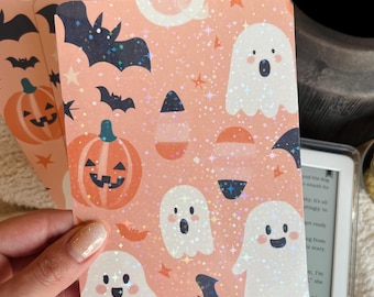 May include: A stack of Halloween-themed cards with a peach background. The cards feature illustrations of ghosts, bats, pumpkins, and candy corn. The designs have a glittery, holographic effect. The cards are held in a hand.