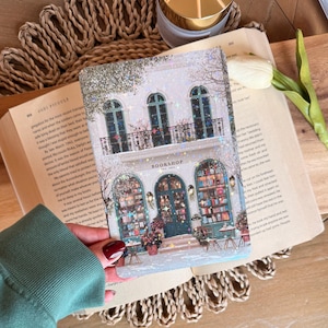 May include: A decorative notebook featuring a detailed illustration of a bookstore facade with arched windows, a teal door, and the text "BOOKSHOP EST. 2023". The design includes floral accents and a snowy effect.