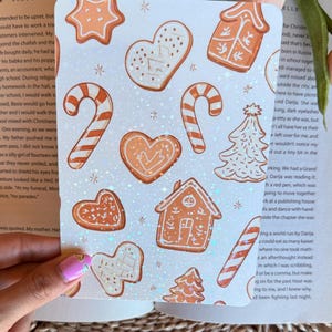 May include: A sheet of stickers featuring various gingerbread cookie designs. The stickers include gingerbread men, hearts, stars, candy canes, and houses, all in shades of brown and white with glitter accents. The stickers are on a white background.