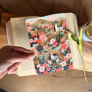 May include: A rectangular art print featuring a vibrant landscape illustration with rolling hills, small white houses, and dense clusters of pink, orange, and green trees. The foreground is filled with detailed floral patterns in shades of pink and red.