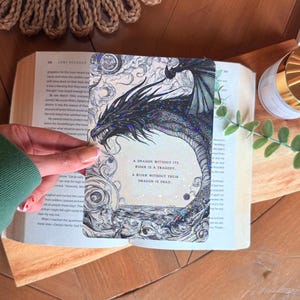 May include: A bookmark featuring a detailed illustration of a black dragon with iridescent accents. The bookmark has text that reads "A dragon without its rider is a tragedy. A rider without their dragon is dead."