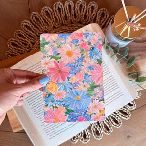 May include: A rectangular art print displaying a profusion of pink, blue, and white flowers. The print is held above an open book, with a woven mat and a gold diffuser in the background. The artwork has a colourful, painterly style, reminiscent of a garden.