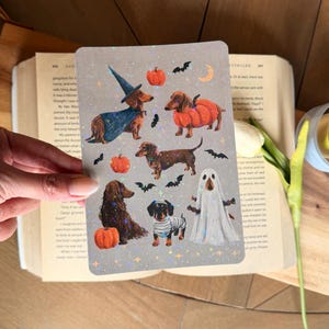 May include: A rectangular card featuring a collection of illustrations of dachshunds in Halloween costumes, including a witch, a pumpkin, a mummy, and a ghost, interspersed with small pumpkins, bats, and holographic stars on a gray background.