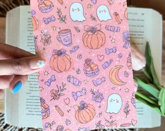 May include: A pink card featuring a pattern of ghosts, pumpkins, sweets, and floral designs. The card is held in front of an open book and a woven placemat.
