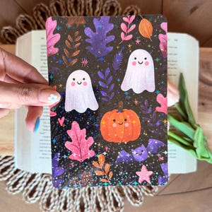 Könnte beinhalten: Eine rechteckige Karte zum Thema Halloween mit schwarzem Hintergrund, verziert mit Blättern, Sternen und Glitzer. Die Karte zeigt Illustrationen von zwei weißen Geistern, einem orangefarbenen Kürbis und einer lila Fledermaus. Die Karte wird von einer Person gehalten.