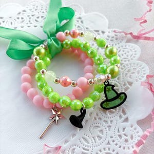 Peut inclure: Trois bracelets superposés avec des perles vertes et roses et des breloques. Les breloques comprennent un chapeau de sorcière, une étoile et une baguette.
