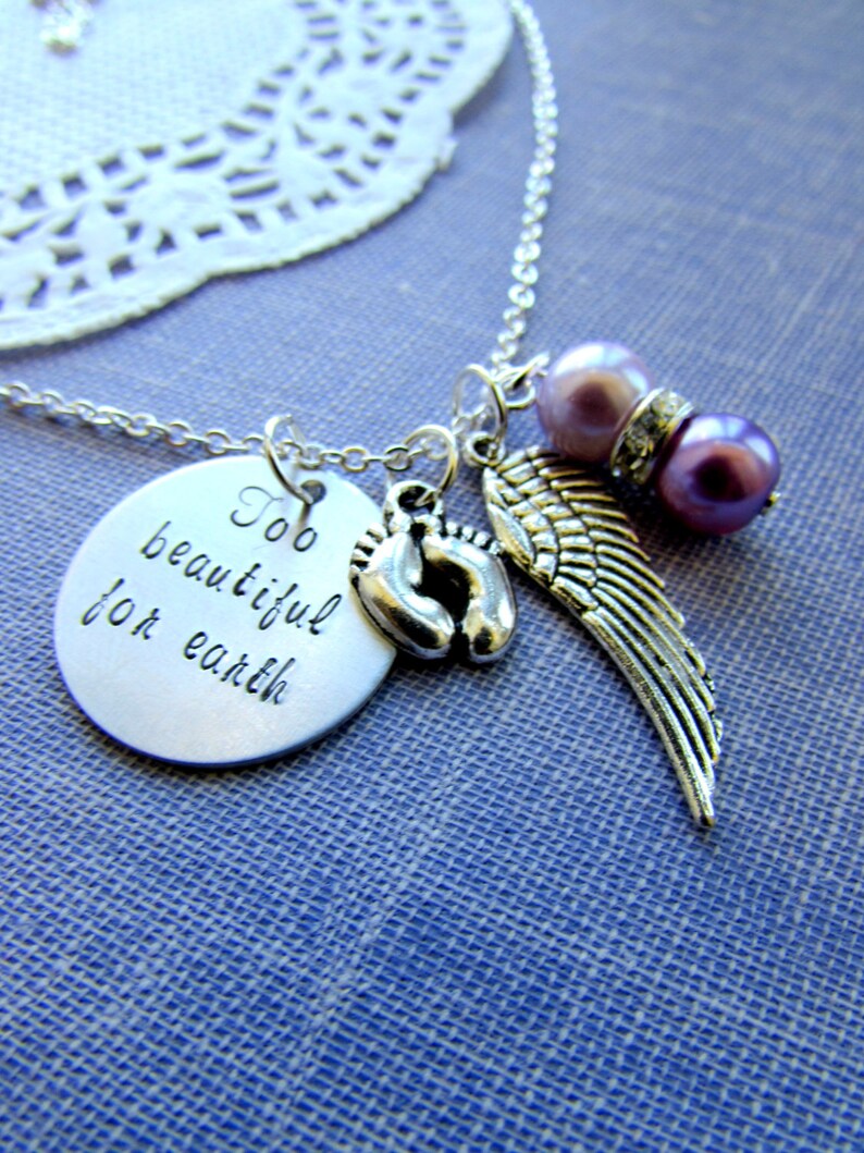 May include: Silver necklace with a charm that reads "Too beautiful for earth", a pair of baby footprints, and a silver angel wing charm. The necklace also features two purple beads.