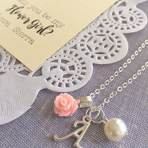 May include: A silver necklace with a pink flower charm, a pearl charm, and a letter A charm. The necklace is on a white lace background with a card that says "Will you be my Flower Girl? Love Sierra".