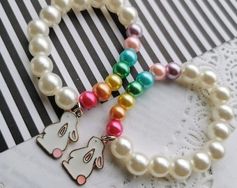 May include: Two white pearl bracelets with rainbow colored beads and a white bunny charm on each. The bracelets are on a white lace doily.