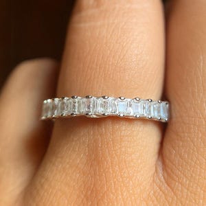 Op de afbeelding: Een zilveren ring met een doorlopende band van rechthoekige, heldere edelstenen. De ring wordt om een vinger gedragen, met een wazige achtergrond.