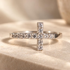 Puede incluir: Un anillo de tono plateado con un diseño de cruz horizontal engastado con múltiples piedras pequeñas, transparentes y de corte redondo. La banda es delgada y pulida, con el motivo de cruz centrado en la parte superior, incrustado con piedras blancas brillantes.