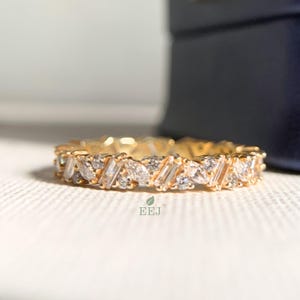 Puede incluir: Un anillo dorado con un patrón repetido de piedras preciosas claras y brillantes. El anillo presenta una mezcla de formas, incluyendo piedras de talla baguette y redondas, colocadas juntas. El anillo está sobre una superficie blanca.