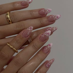 May include: Close-up of long, almond-shaped nails with a clear base and a white and pink floral design. The nails are adorned with small, delicate flowers. Several gold rings are visible on the fingers.