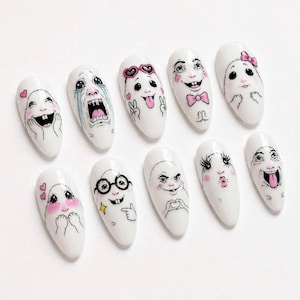 May include: A set of ten white artificial nails, each hand-painted with a different cartoon face. The faces display various expressions, including crying, laughing, and sticking out the tongue. Pink accents are used for cheeks and other details.
