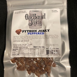 May include: A silver resealable bag containing 1 ounce (28 grams) of Oak Barrel Jerky Python Jerky Peppered. The bag has a label with the company logo and nutritional information. The jerky is a dark brown color and appears to be made from python meat.