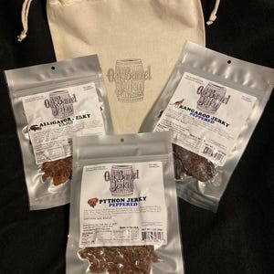 May include: An assortment of jerky products from Oak Barrel Jerky, including alligator, kangaroo, and python varieties. The jerky is packaged in clear plastic bags, with a canvas drawstring bag in the background. The bags feature the Oak Barrel Jerky logo and product information.