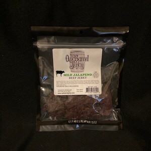 May include: A clear plastic bag containing a 3 oz bag of Oak Barrel Jerky Mild Jalapeno Beef Jerky. The bag is sealed and has a white label with the company logo and product information. The jerky is dark brown in color and appears to be sliced into thin strips.