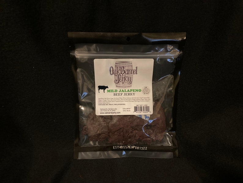 May include: A clear plastic bag containing a 3 oz bag of Oak Barrel Jerky Mild Jalapeno Beef Jerky. The bag is sealed and has a white label with the company logo and product information. The jerky is dark brown in color and appears to be sliced into thin strips.