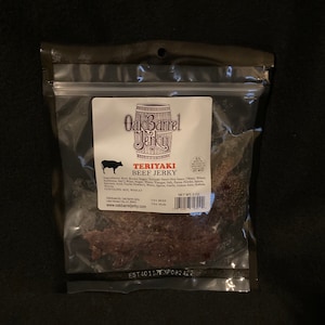 May include: A clear plastic bag with a resealable zipper closure contains a package of Oak Barrel Jerky Teriyaki Beef Jerky. The package is labeled with the company name and logo, and the ingredients and nutritional information are listed on the back. The jerky is a dark brown color and has a slightly textured surface. The bag is on a dark brown background.