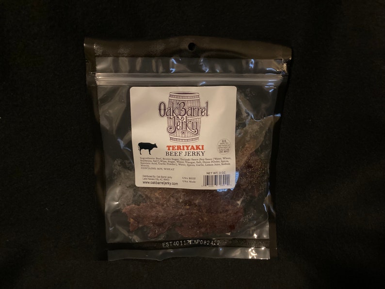May include: A clear plastic bag with a resealable zipper closure contains a package of Oak Barrel Jerky Teriyaki Beef Jerky. The package is labeled with the company name and logo, and the ingredients and nutritional information are listed on the back. The jerky is a dark brown color and has a slightly textured surface. The bag is on a dark brown background.