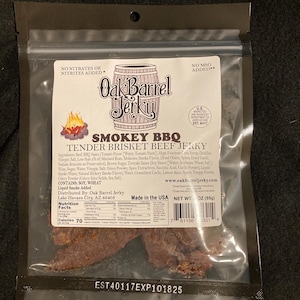 May include: A resealable bag of Oak Barrel Jerky, Smokey BBQ Tender Brisket Beef Jerky. The bag is open to reveal three pieces of jerky. The bag is labeled with the company's logo and information about the product. The bag is made in the USA and has a net weight of 3 oz (85g).