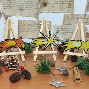 Op de afbeelding: Drie geborduurde komeetpatches in oranje, geel en wit worden tentoongesteld op kleine houten ezels. De patches zijn omgeven door dennenappels, groen en decoratieve elementen.