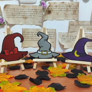 Può includere: Tre cappelli da strega decorativi in bordeaux, grigio e viola, ciascuno su un cavalletto di legno in miniatura. Il cappello bordeaux ha un fiocco rosso, il cappello grigio ha una fibbia dorata e il cappello viola ha una luna crescente gialla. Foglie autunnali e ritagli neri circondano i cappelli.