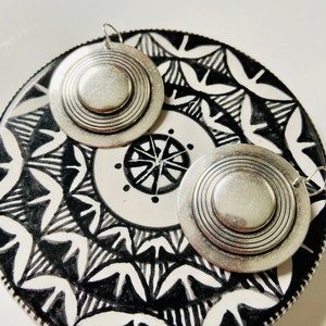 May include: A pair of silver earrings with a circular design. The earrings have a layered, concentric circle pattern.