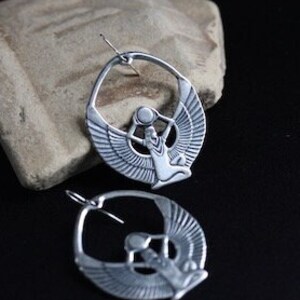May include: A pair of silver hoop earrings featuring a detailed cutout design of a winged figure holding a sphere. The earrings are hanging from a rough, textured surface.